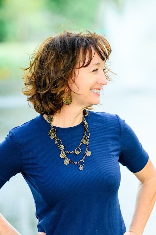 Smiling woman in a blue t-shirt with wavy hair and decorative necklace, standing outdoors with a blurred background.